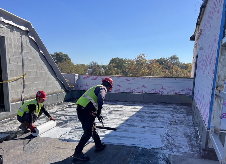 Two roofing workers in safety gear using brooms to spread waterproofing roofing material